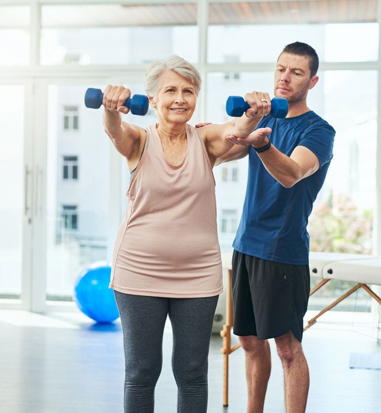 Senior woman lifting dumbbells with the assistance of a trainer in a bright living unit gym.
