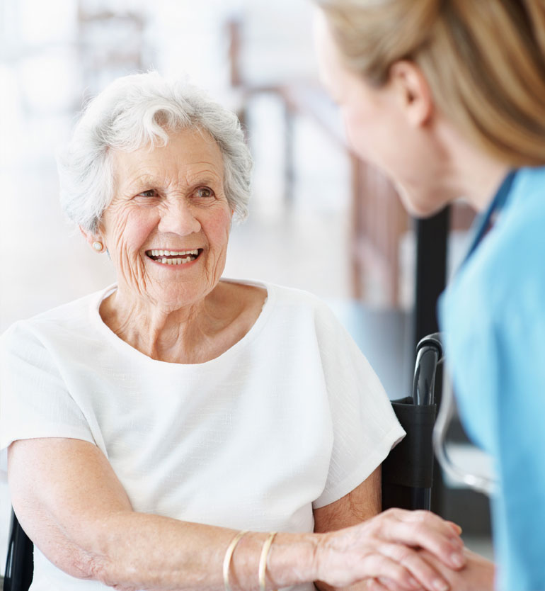 Elderly woman in a white shirt smiling at a caregiver in a senior living unit.