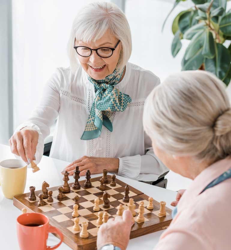 Two elderly women enjoy a game of chess at a senior living community, smiling and drinking coffee.