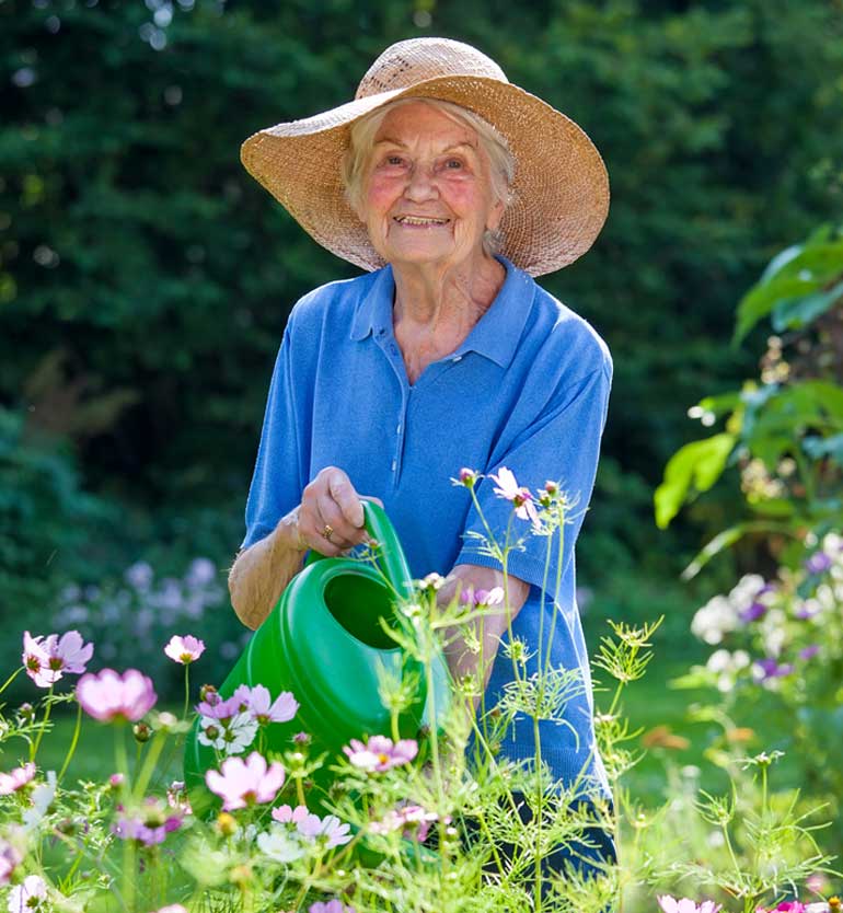 Elderly woman in a sunhat watering flowers in a garden at a senior living community.