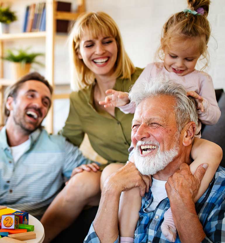Elderly man laughing with family, including a child on his shoulders, in a cozy living room.