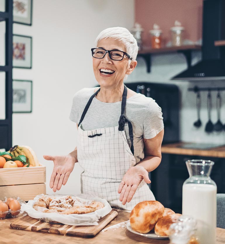 Senior woman smiling and presenting freshly baked goods in a modern kitchen at a senior living unit.