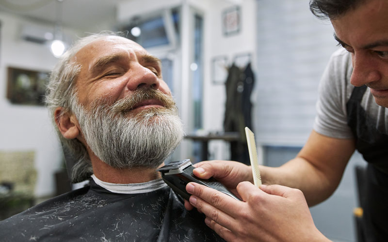 Senior man enjoying a beard trim by a barber in a well-lit salon.