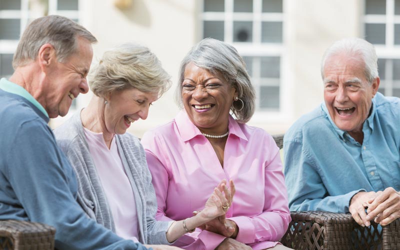 Four seniors laughing together outside a senior living community building on a sunny day.
